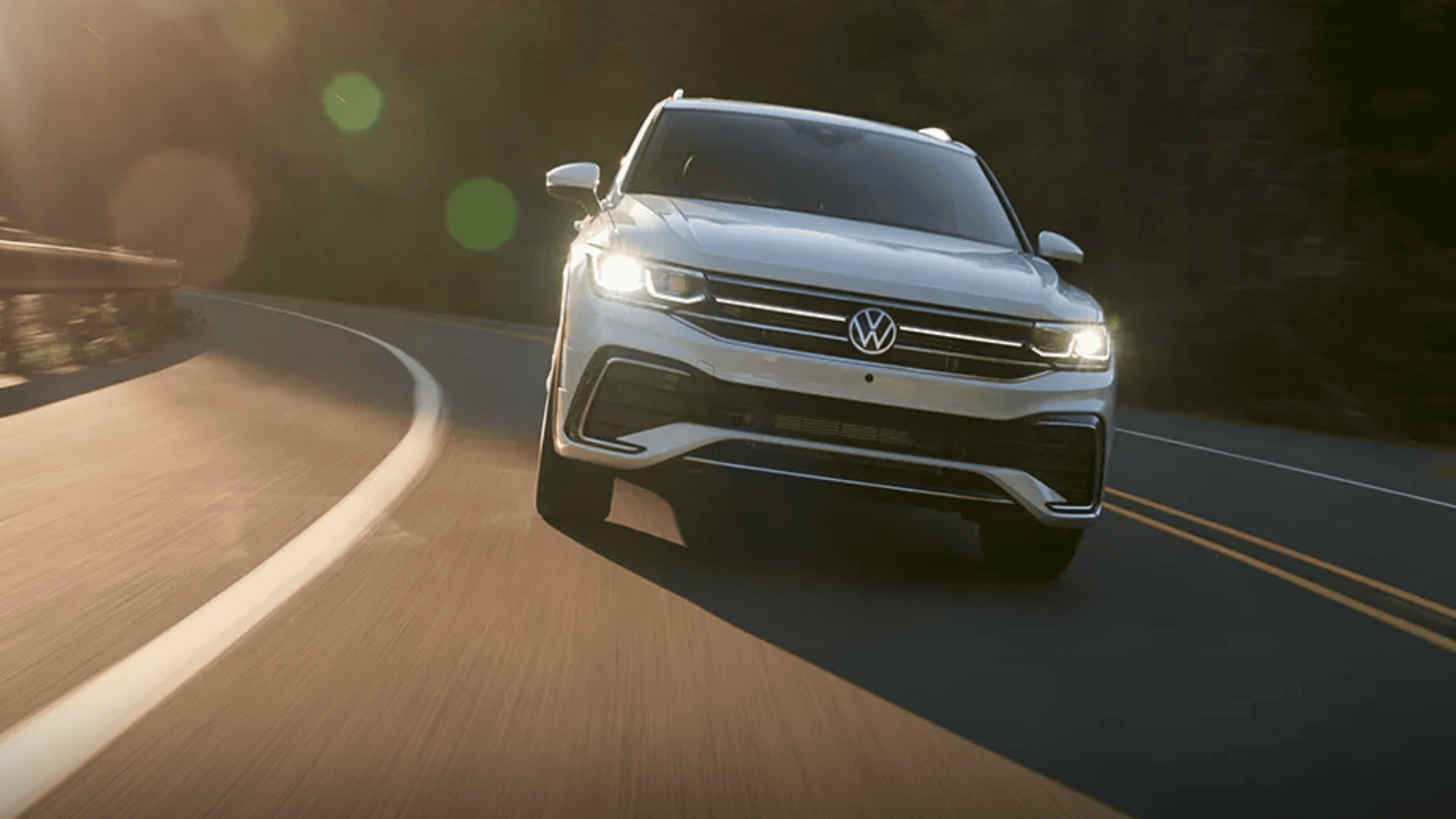 2024 Volkswagen Tiguan SUV driving on a curved mountain road with headlights on, showcasing its sleek front grille and modern design in natural sunlight.
