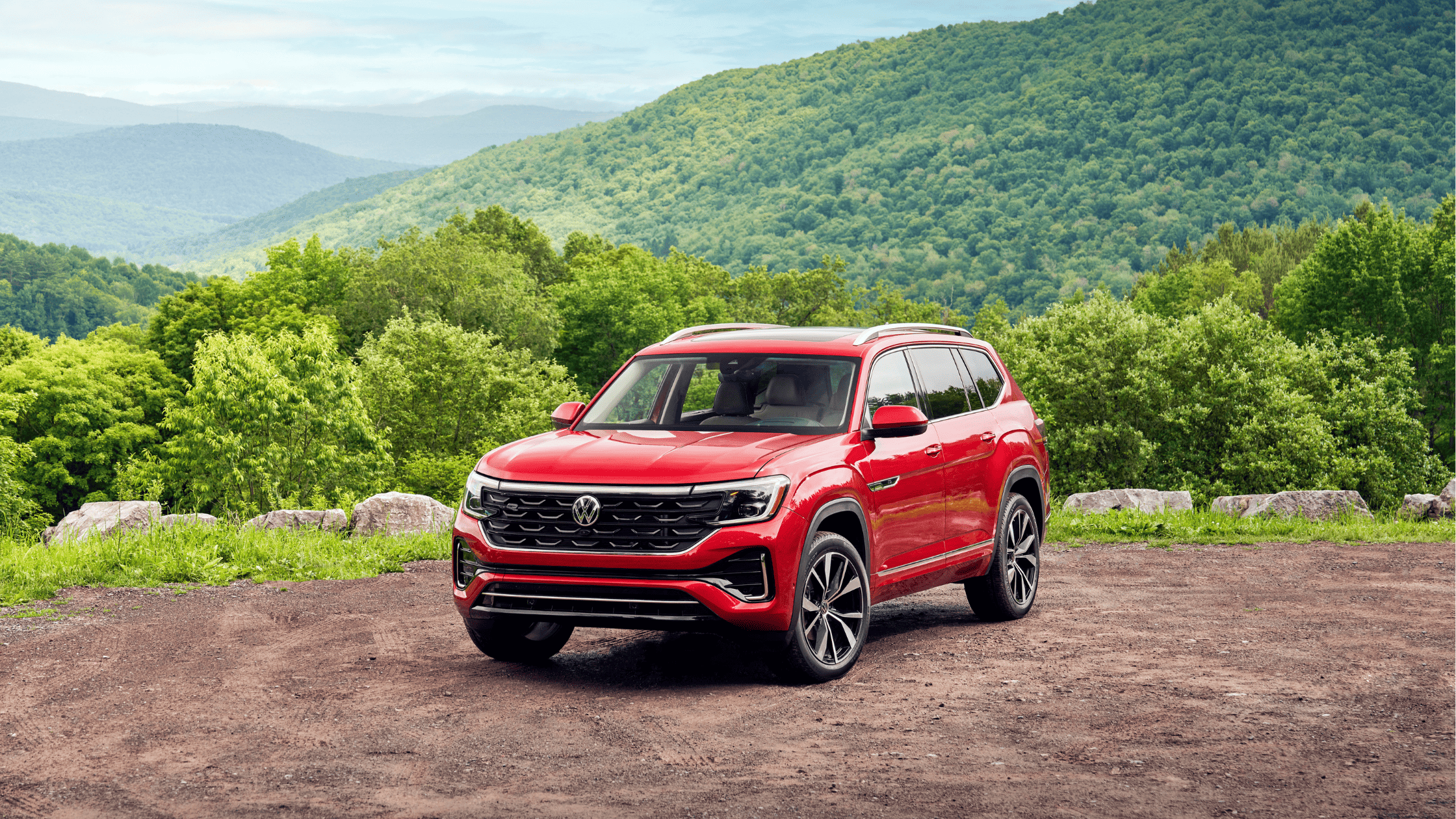 2024 Volkswagen Atlas SUV in bold red parked on a mountain overlook with lush green hills and a scenic valley in the background, emphasizing its rugged design and spaciousness for adventure.