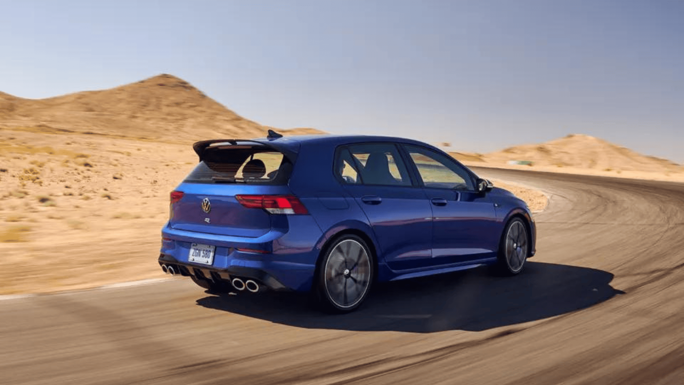 2024 Volkswagen Golf R in vibrant blue driving on a desert racetrack, highlighting the car’s sporty rear design, quad exhaust, and performance features in an open landscape.