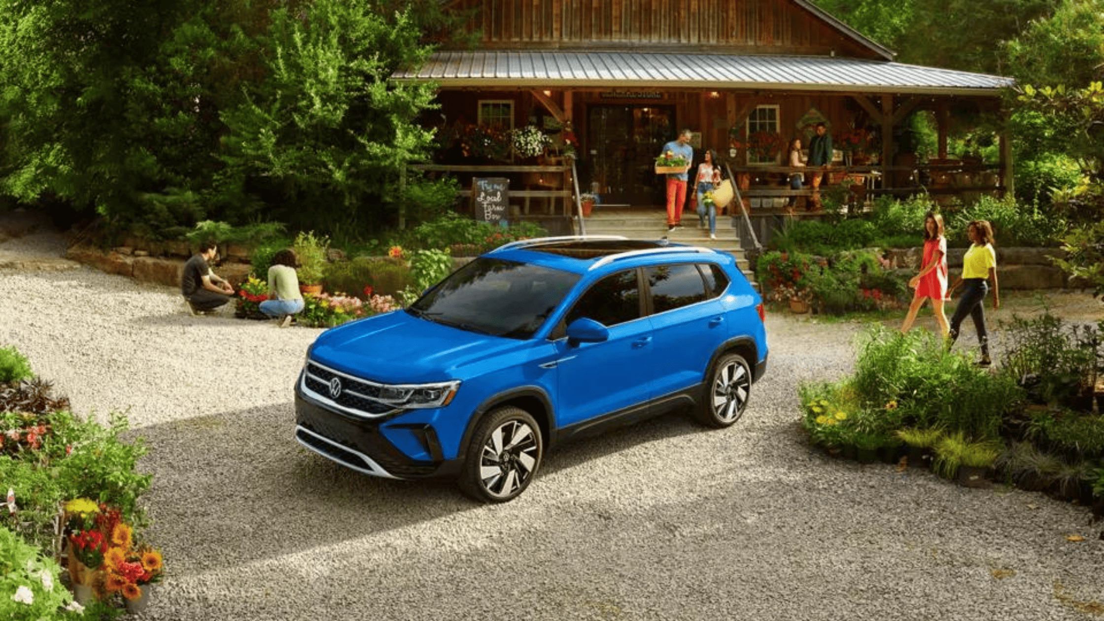 2024 Volkswagen Taos SUV in bright blue parked in front of a rustic wooden store with people and garden plants, highlighting the vehicle’s stylish design and outdoor lifestyle appeal.