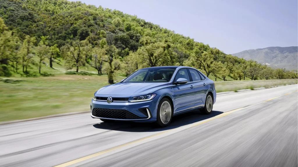 A blue 2025 Volkswagen Jetta driving on a scenic highway with greenery.
