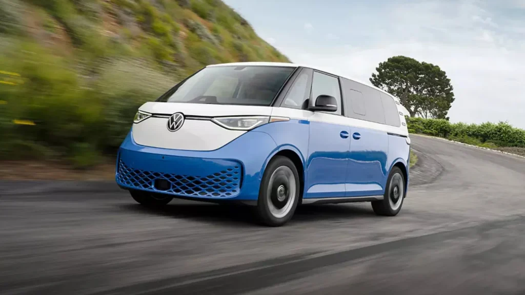 Blue and white 2025 Volkswagen ID. Buzz driving around a curve in scenic terrain.