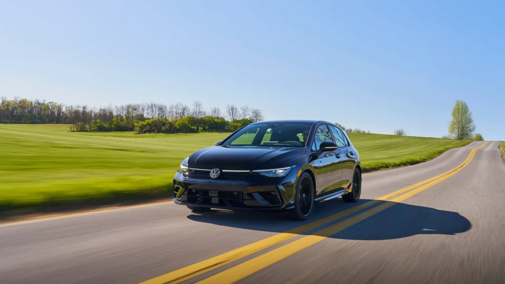 2025 Volkswagen Golf R in black driving on a winding rural road under clear blue skies.