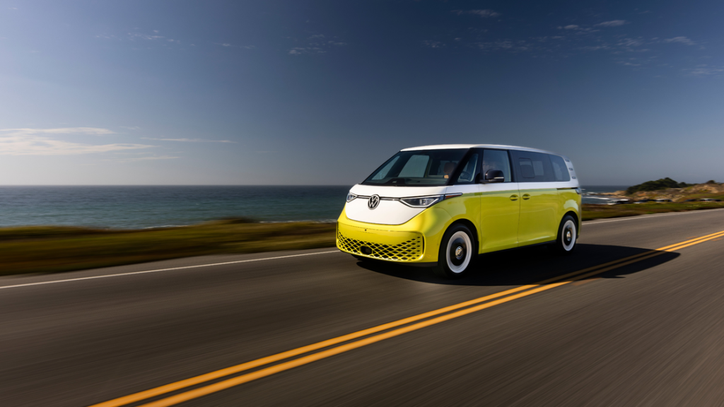 Front-angle view of a two-tone yellow and white 2025 Volkswagen ID. Buzz driving along a coastal highway.