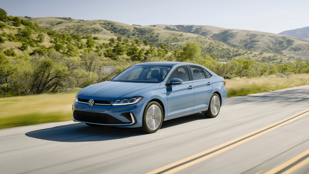 Blue 2025 Volkswagen Jetta sedan driving on open highway with scenic hills in the background.