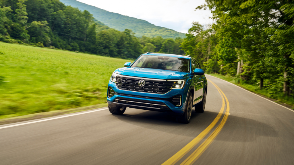 2026 Volkswagen Atlas Cross Sport SUV driving on a winding road with green hills and mountain scenery.