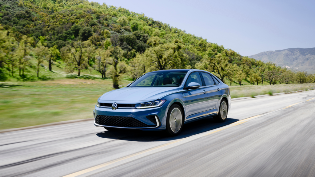 2026 Volkswagen Jetta sedan driving on an open highway with trees and rolling hills in the background.
