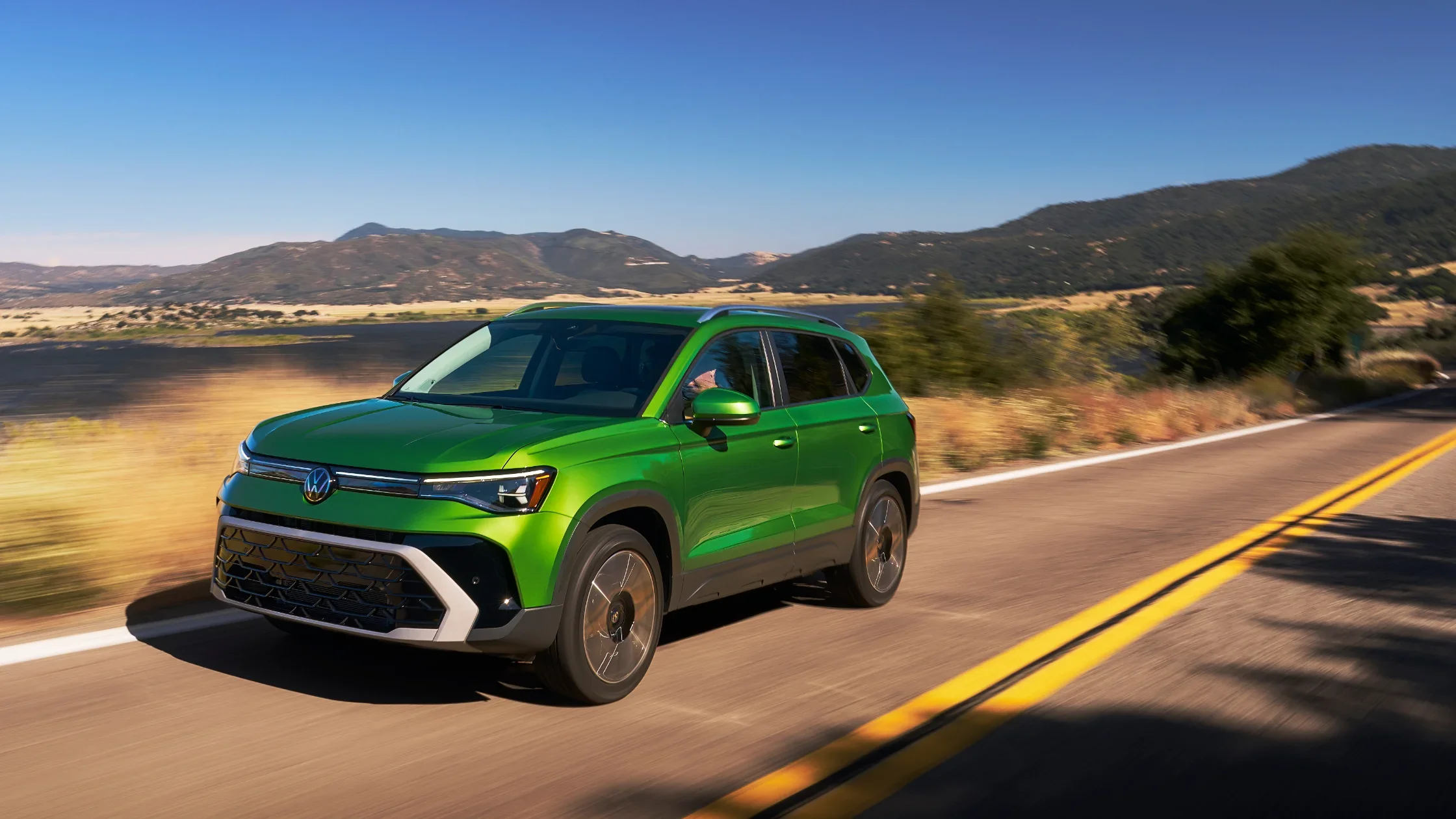 2025 Volkswagen Taos in green driving on a scenic highway with trees and hills in the background.
