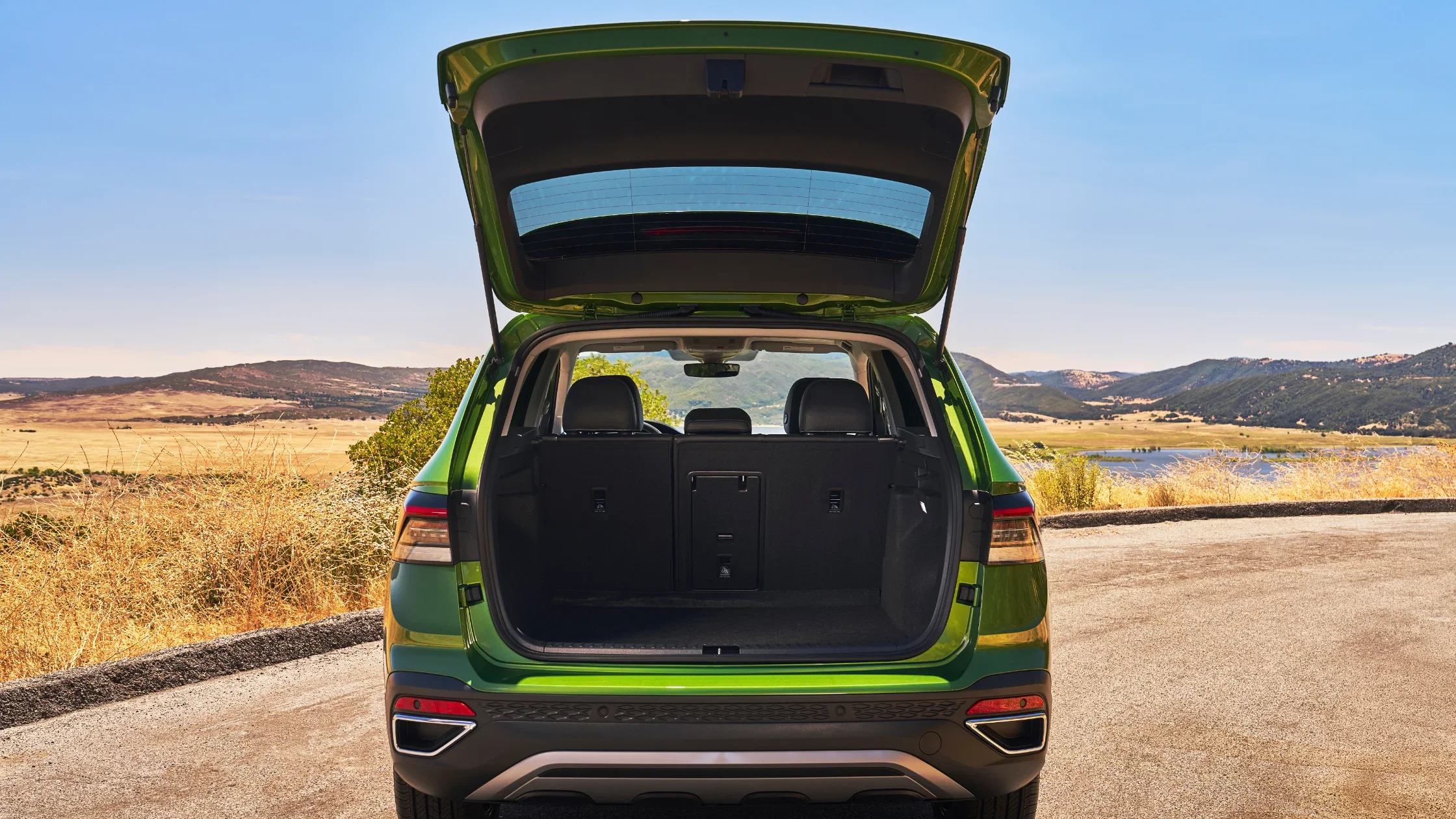 Rear view of the 2025 Volkswagen Taos with the tailgate open, displaying spacious cargo area against a scenic mountain and lake backdrop.
