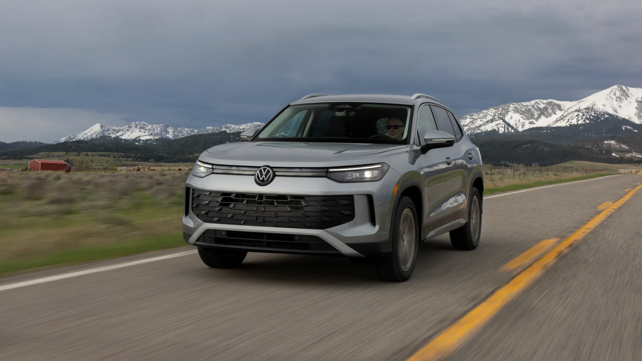 Silver 2025 Volkswagen Tiguan driving on a scenic road with snowy mountains in the background.