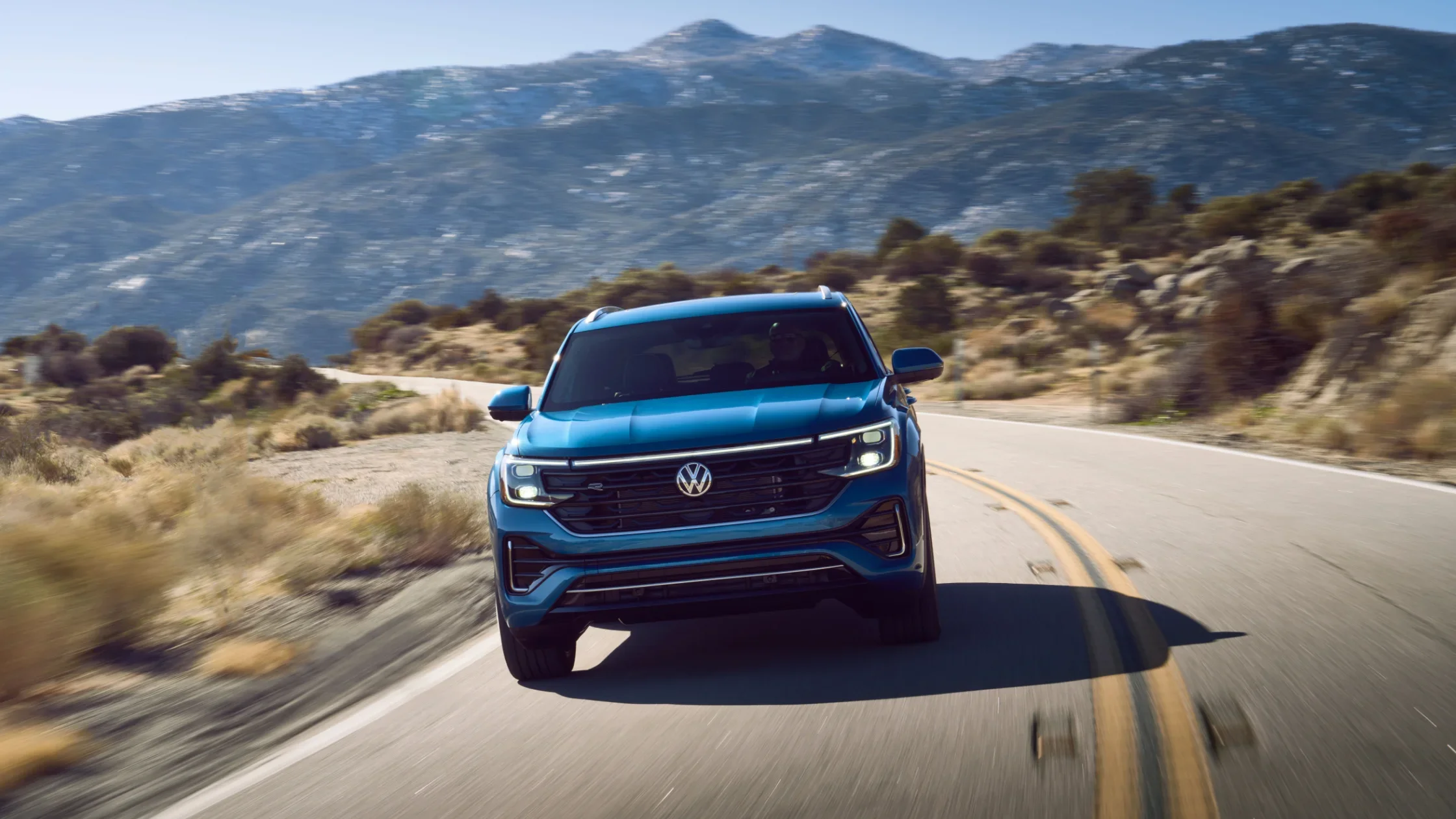 Front view of a blue 2025 Volkswagen Atlas Cross Sport driving on a winding mountain road under clear skies.