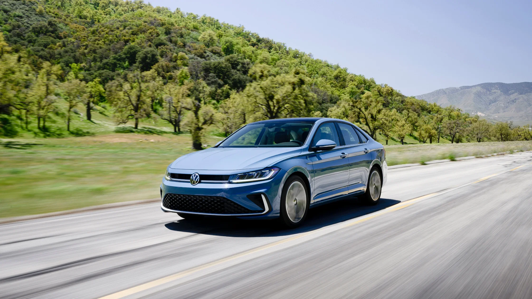 2025 Volkswagen Jetta in motion on a countryside road, surrounded by green trees and distant mountains under a clear blue sky.