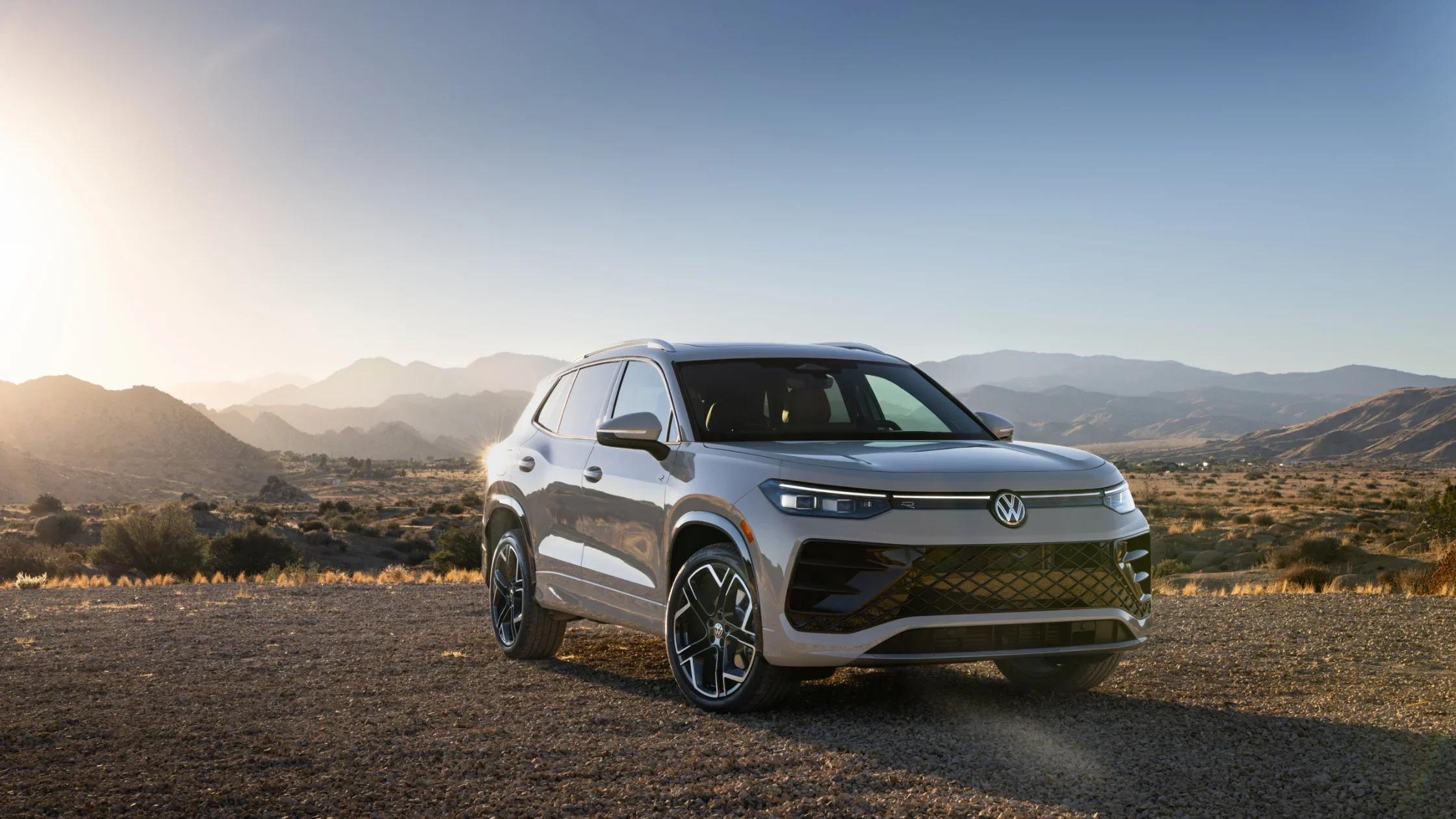 2025 Volkswagen Tiguan SUV parked in a desert landscape at sunset, showcasing its sleek design and silver finish against a backdrop of distant mountains.