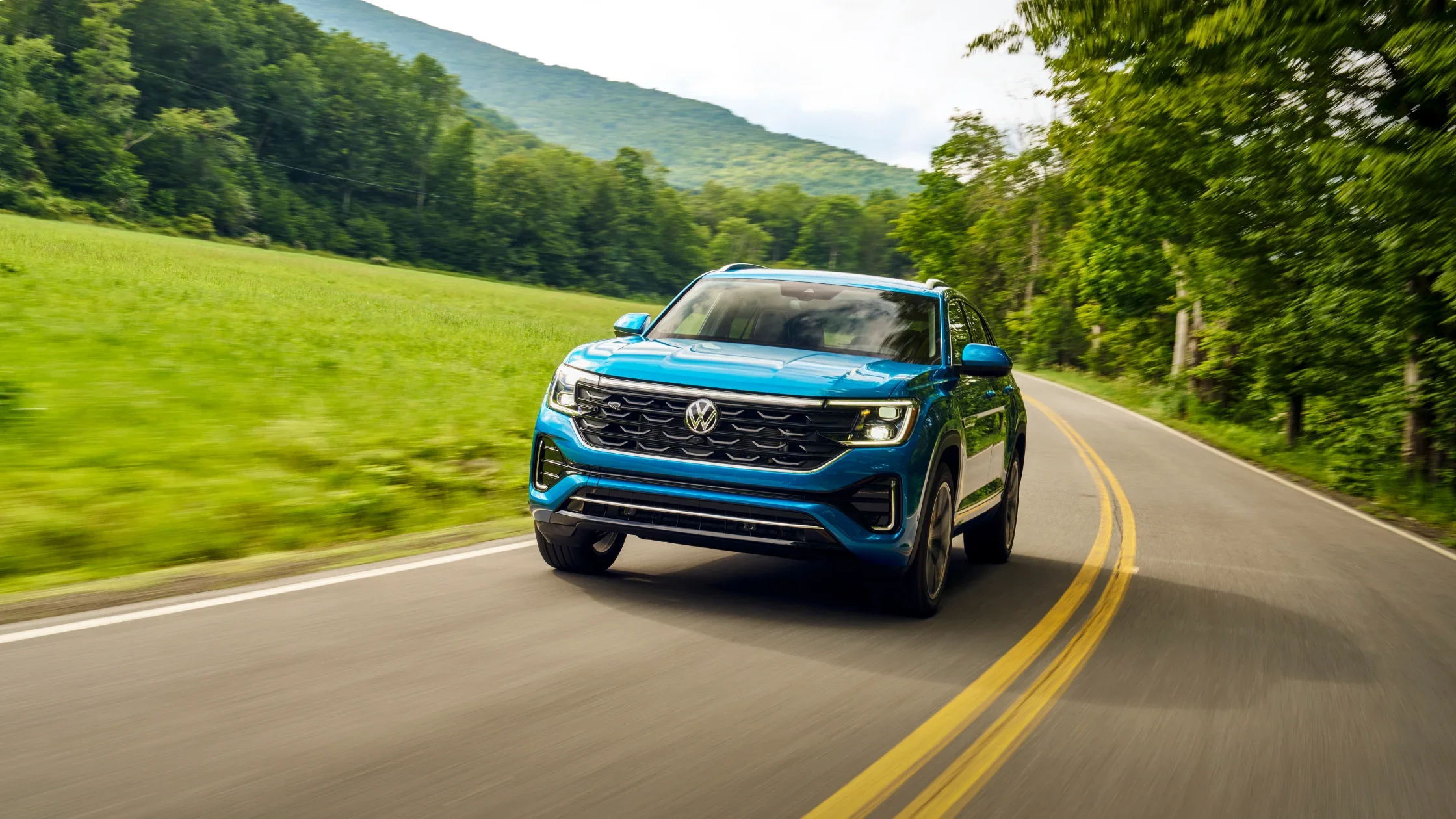 2026 Volkswagen Atlas Cross Sport exterior front view driving on scenic road in blue.