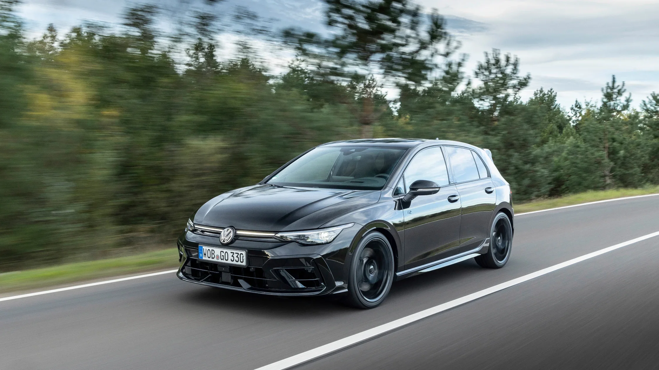 Front three-quarter view of the 2025 Volkswagen Golf R in motion, highlighting its aggressive front fascia, blacked-out wheels, and high-performance handling on a scenic road.