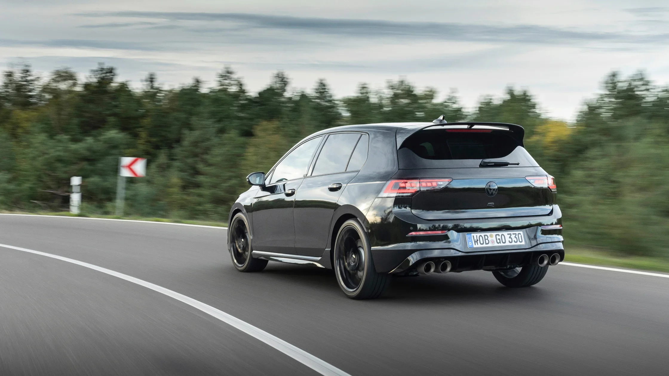 Rear view of the 2025 Volkswagen Golf R in sleek black, driving on a winding road, showcasing its quad exhaust system, sporty stance, and aerodynamic design.