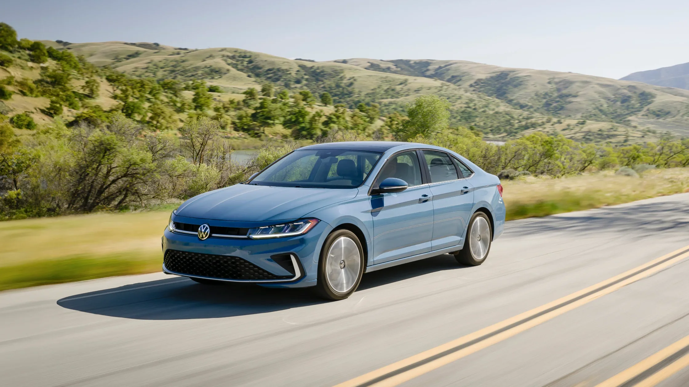 Blue 2025 Volkswagen Jetta driving on a scenic road with rolling hills.