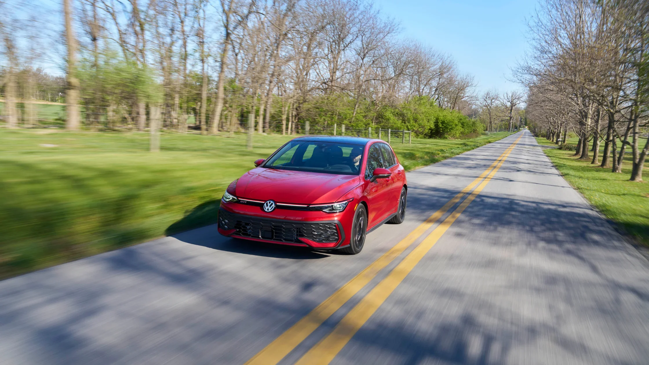 Red 2025 Volkswagen Golf GTI driving down a tree-lined road with motion blur effect.