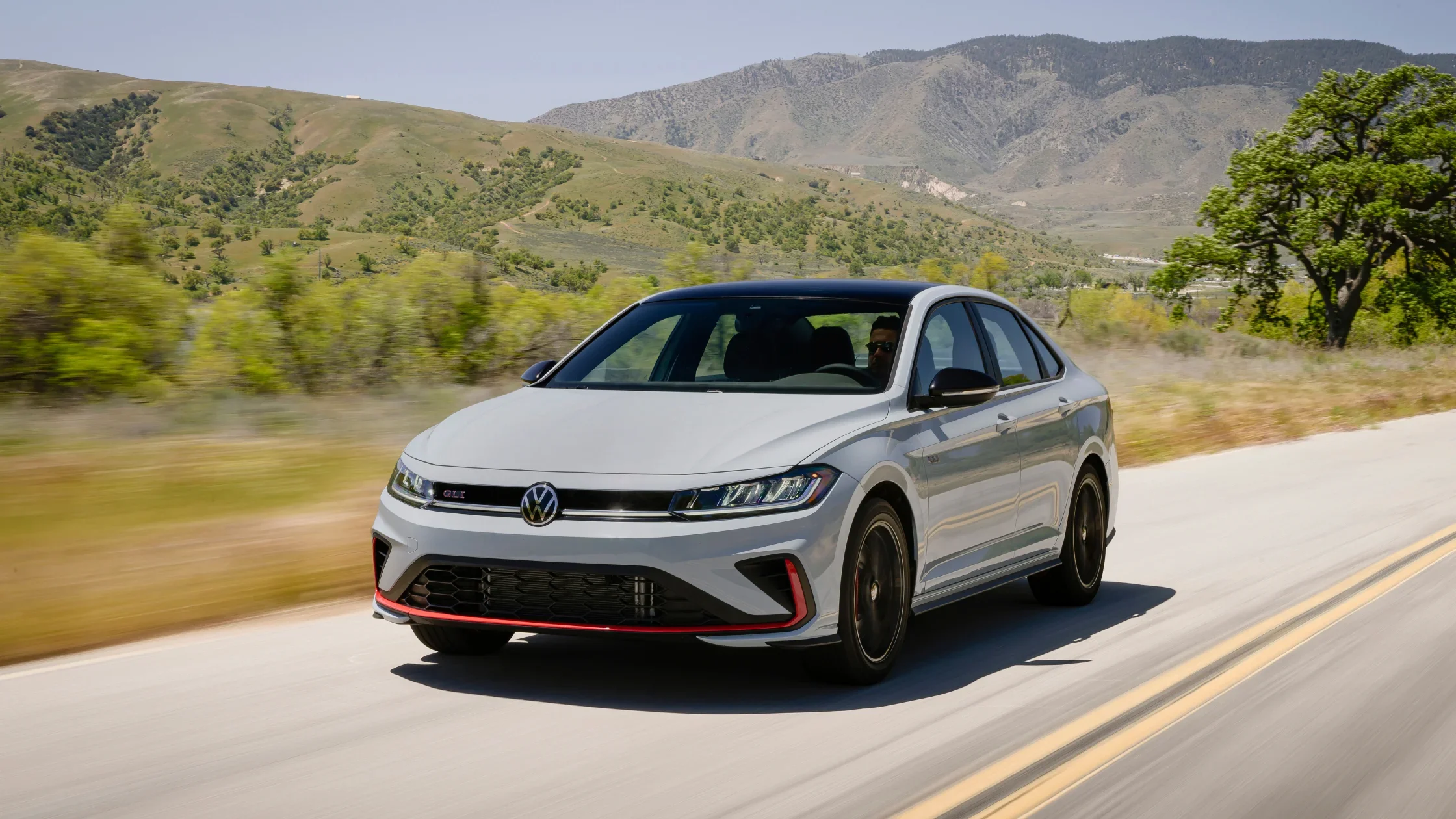 2026 Volkswagen Jetta GLI white sport sedan driving on a mountain road in front three quarter view.