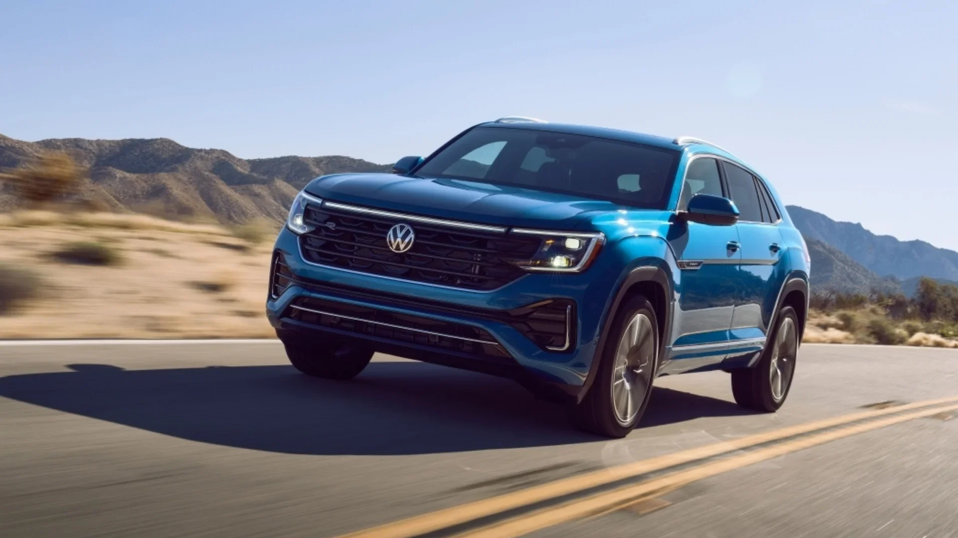Blue 2026 Volkswagen Atlas Cross Sport driving on a desert road with mountains in the background.